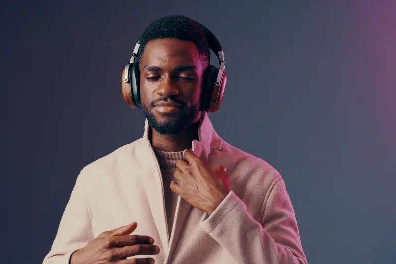 A stylish young Black man enjoying music with headphones, wearing a cozy beige jacket against a colorful gradient background, evoking relaxation and joy