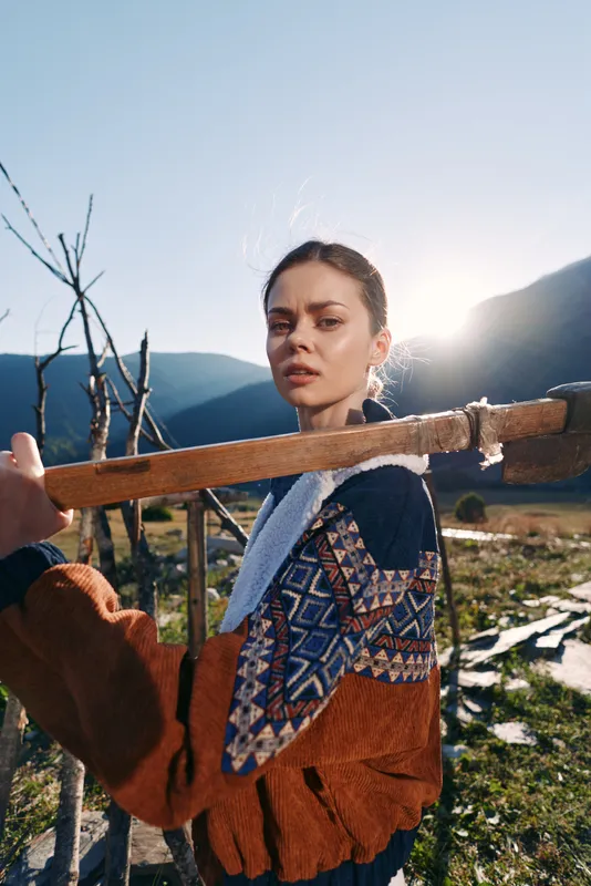 Woman portrait outdoor in mountains with sunlight wearing patterned sweater and holding wooden stick in rural landscape, nature scene with thoughtful expression and travel vibe