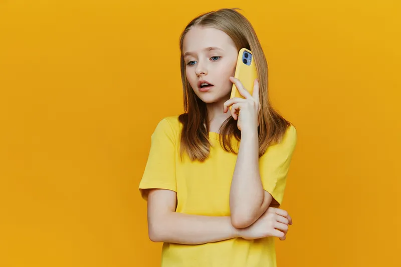 Young girl in a yellow shirt talking on a yellow phone against a bright yellow background, conveying emotions of surprise and curiosity