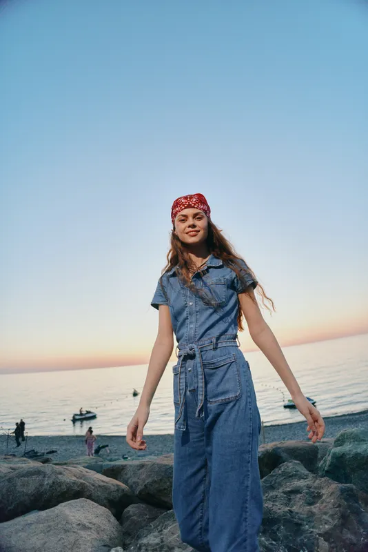 smiling woman in denim dress with red bandana standing on rocks by the ocean during sunset with calm sea and clear sky