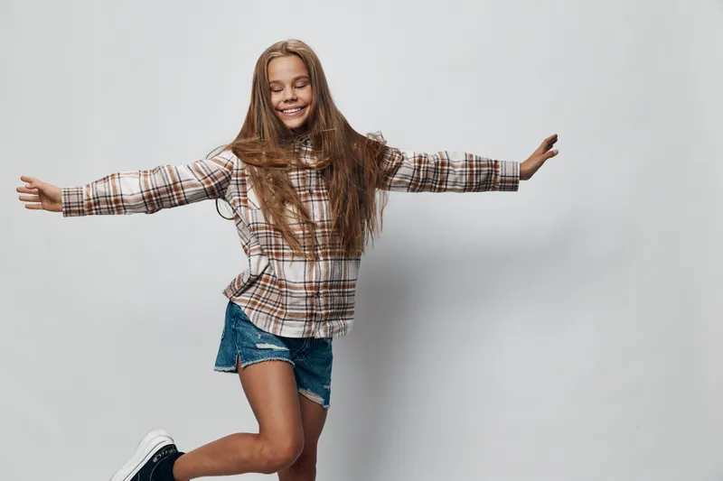 Girl jumping joyfully with eyes closed and arms outstretched wearing plaid shirt and denim shorts on white background with happy smile and long hair in motion.