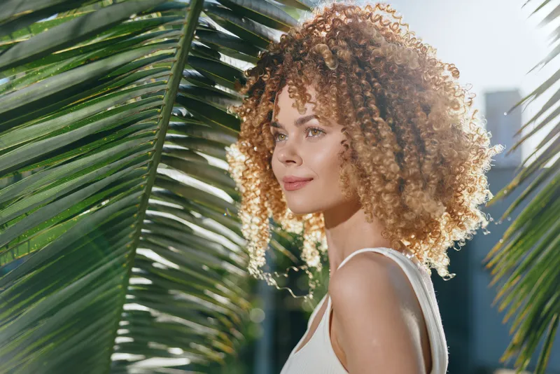 Curly woman with natural hair standing near palm leaves in bright sunlight, wearing white sleeveless top, looking serene and radiant outdoors.