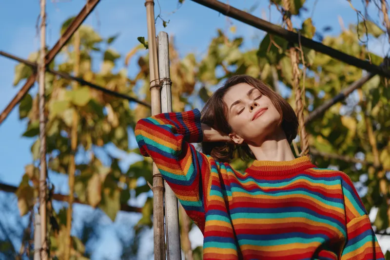 Woman in her twenties wearing a colorful striped sweater, leaning on a wooden trellis, smiling with eyes closed in a sunny outdoor garden, relaxed lifestyle and fashion portrait.