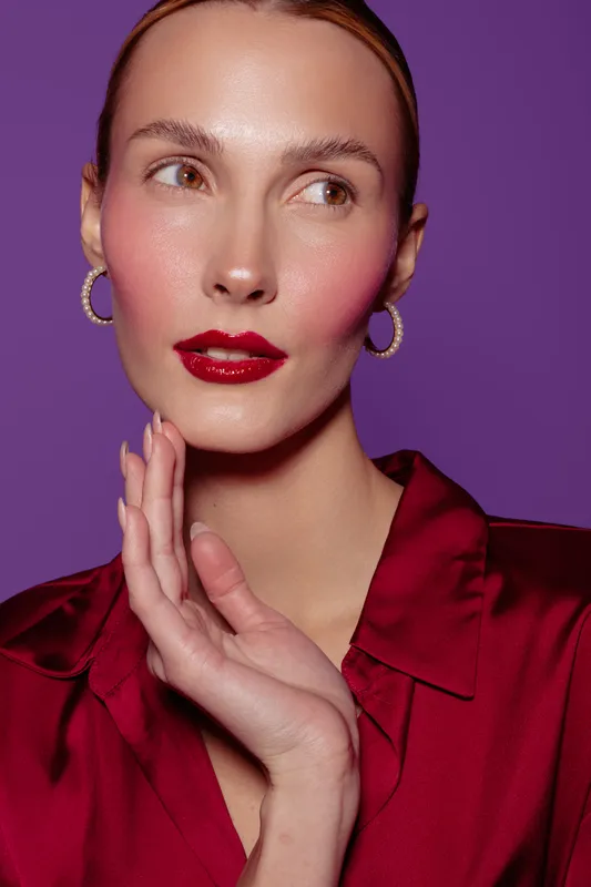 Portrait of a woman with glossy red lips and pink blush wearing hoop earrings and a red satin blouse. She poses against a purple background with soft lighting and a thoughtful expression.