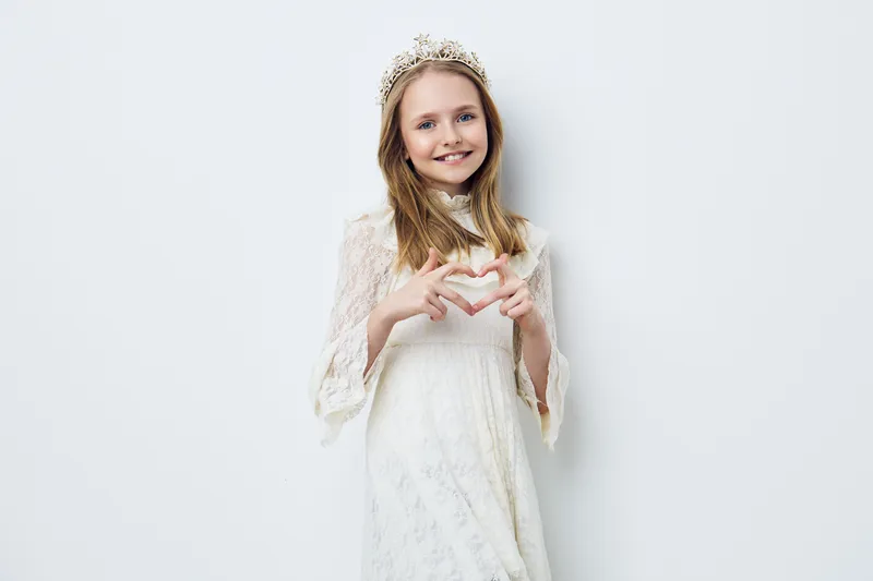 Young girl in a white dress and crown making heart shape with hands, joyful expression, minimalist white background, capturing innocence and beauty