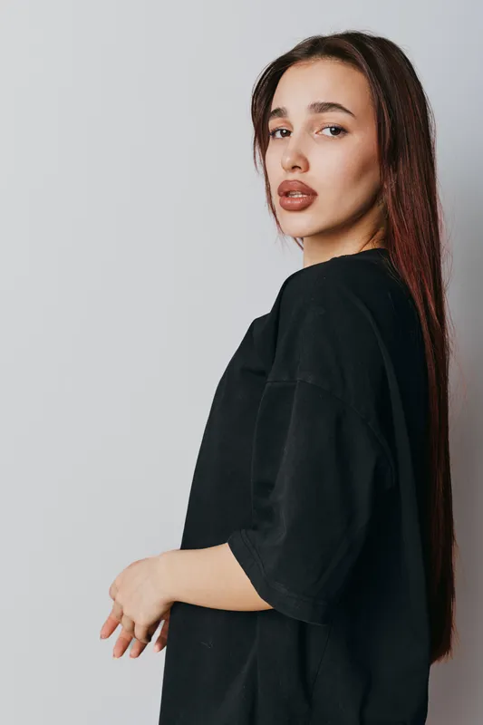 Young woman with long brown hair and natural makeup posing in black oversized shirt against white background. Casual style, confident expression and modern fashion portrait.