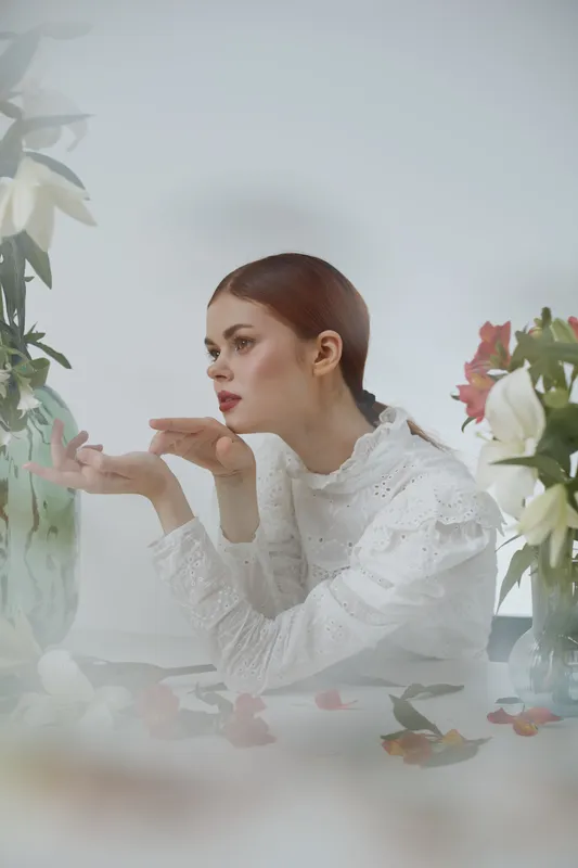 Elegant young woman in white attire expressing contemplation, surrounded by flowers in a soft, ethereal atmosphere, reflecting tranquility and beauty