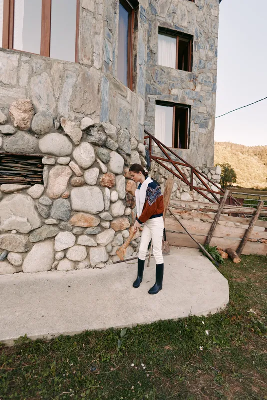 Woman in sweater and boots walking by a stone building in countryside near wooden stairs and rustic facade, casual autumn outfit and relaxed rural lifestyle scene.