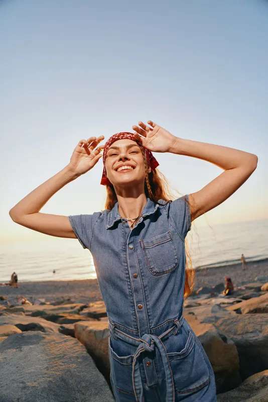 smiling woman in denim jumpsuit enjoying sunny beach day with red headscarf, long hair, warm light and relaxed joyful mood