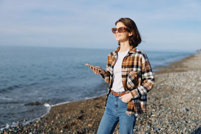 Beach scene with a woman on a sunny shore using a smartphone, wearing a plaid shirt and denim jeans, enjoying daylight by the sea.