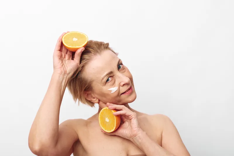 Mature woman with short hair holds two orange halves near her face with white cream spots on cheeks against white background. Concept of natural skincare and healthy lifestyle.