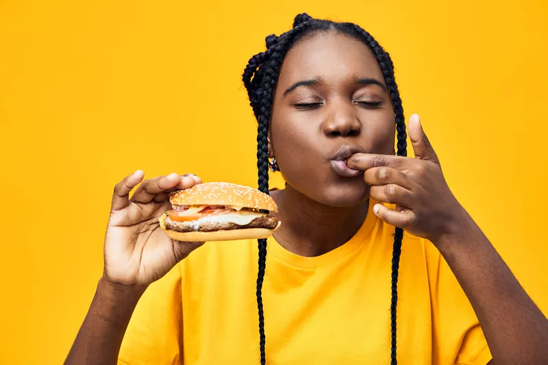 Young woman enjoying a delicious hamburger with a playful expression, vibrant yellow background, emphasizing a fun and indulgent food experience
