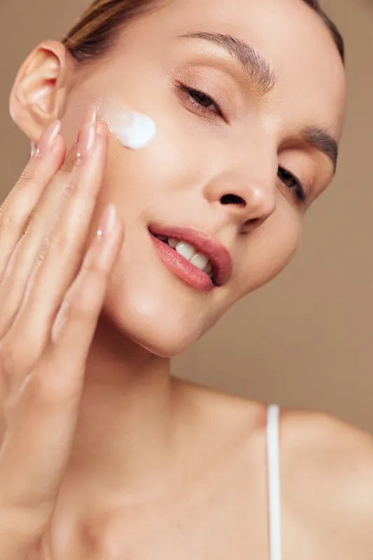 Close up of woman applying face cream with fingers, smooth skin, natural makeup, soft lighting, skincare routine, beige background, gentle expression