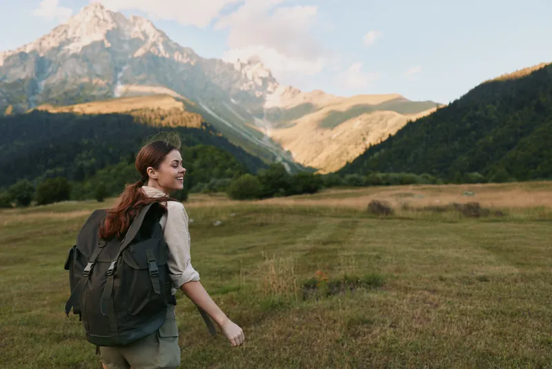 Bright woman hiking outdoors with a backpack, scenic meadow and distant mountains, warm sunlight, relaxed stride, natural colors, sense of adventure