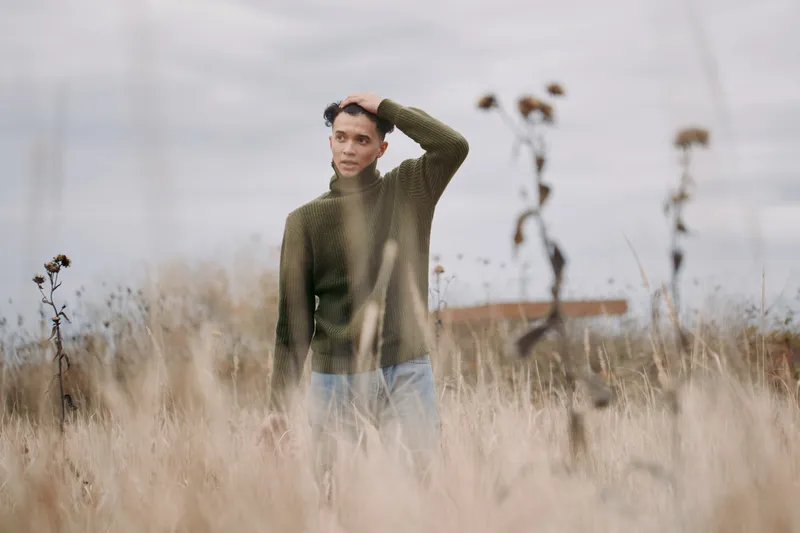 A genuine candid portrait of a young man standing in tall dry grass wearing a knit green sweater, captured with natural light to highlight a sincere expression and realistic outdoor atmosphere.