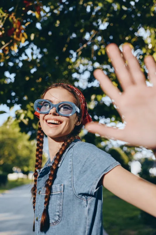 Lifestyle woman with modern boho-western denim street style smiling and wearing blue sunglasses, outdoors with natural green background in vibrant film color.