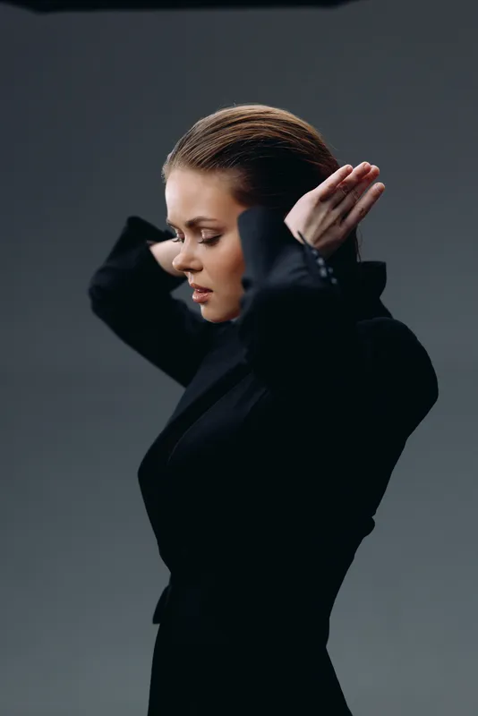 Young woman in black jacket fixing her hair with serious expression on gray background
