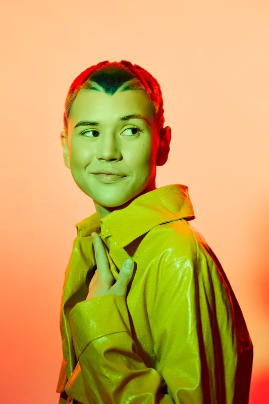 young woman with short colorful hair wearing a yellow leather jacket, looking sideways under vibrant orange and green lighting, modern urban style portrait