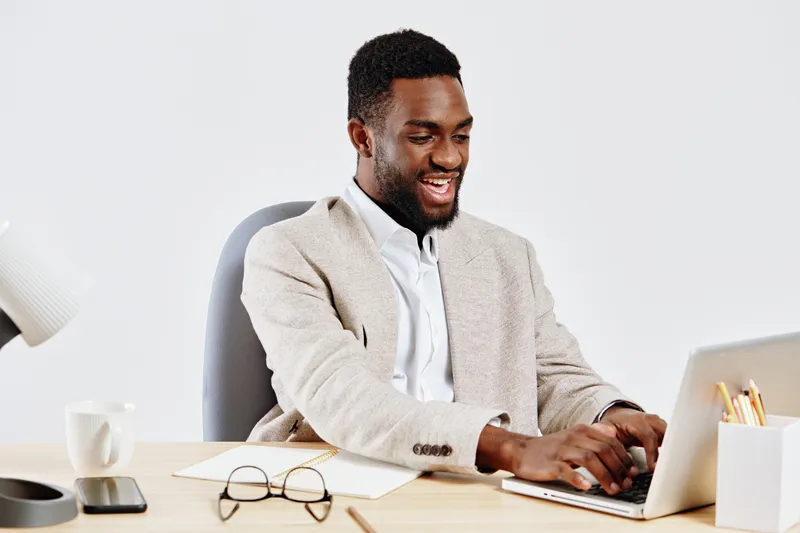 Professional smiling man working on laptop at modern office desk, wearing smart beige blazer and white shirt, with glasses, coffee mug, and notepad, indoors, business casual style, organic natural lig