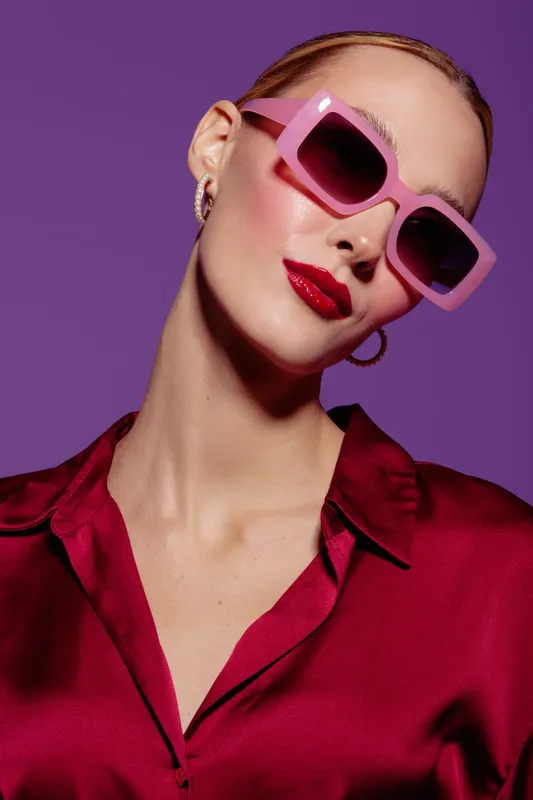 Stylish woman with slicked hair wearing pink sunglasses and red lipstick poses confidently against purple background. Fashion portrait with bold makeup and trendy accessories concept.