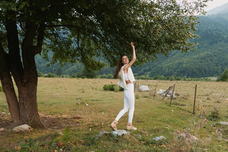 Young woman in white stands beside a sturdy tree in a sunlit meadow, raising one arm in a playful, carefree pose with distant mountains in the background and a rustic fence.