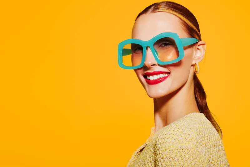 Smiling woman with large turquoise glasses and red lipstick poses against bright yellow background. Fashion portrait with vibrant colors and confident expression. Concept of style and beauty.