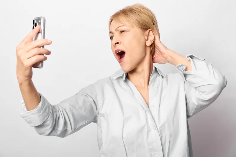Shocked surprised woman holds smartphone for selfie on white background, phone in left hand, touches ear with right hand, listening music loudly, open mouth expression, studio portrait