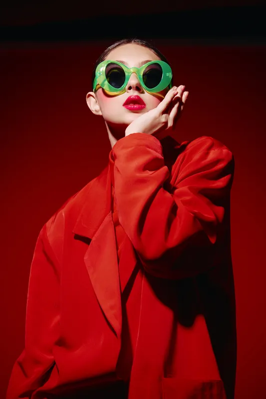 Fashion woman in oversized red blazer poses wearing bold green circular sunglasses and vibrant red lipstick against a glossy red background in a creative beauty concept.