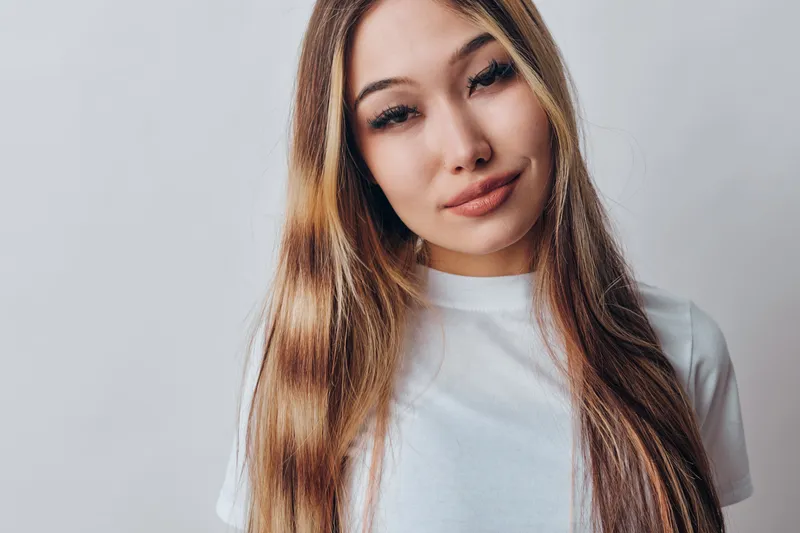 Young woman with long straight hair wearing white t shirt looking at camera with gentle smile and relaxed posture against plain background in studio light.