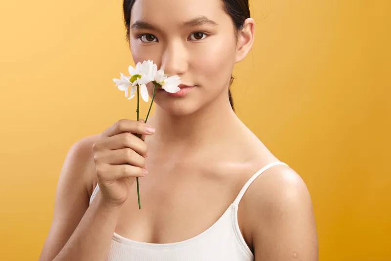 asian woman holds a delicate flower near her face, posing against a warm orange backdrop. soft lighting highlights smooth skin, calm expression, and graceful presence. The composition conveys natural