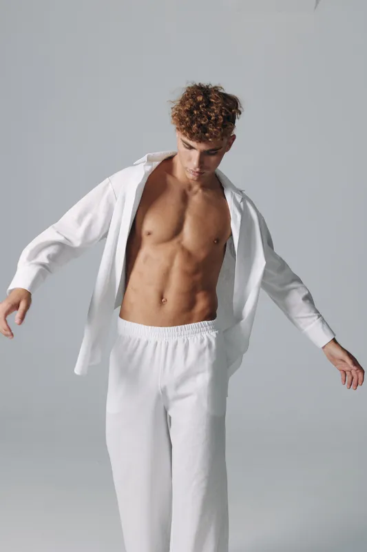 Young man with curly hair wearing white pants and open shirt showing toned muscular torso in studio with neutral background and soft lighting.