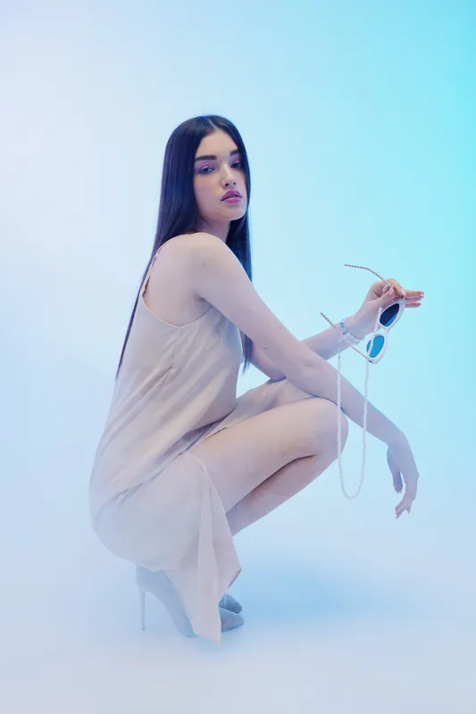Young woman in a light dress squats while holding sunglasses in a soft blue studio light. Concept of beauty, fashion, elegance, and modern style.