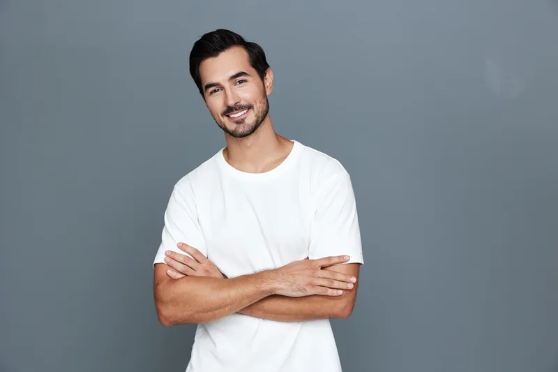 Handsome smiling man wearing a white t-shirt poses confidently with arms crossed against a solid gray background, exuding approachability and style in this studio portrait.