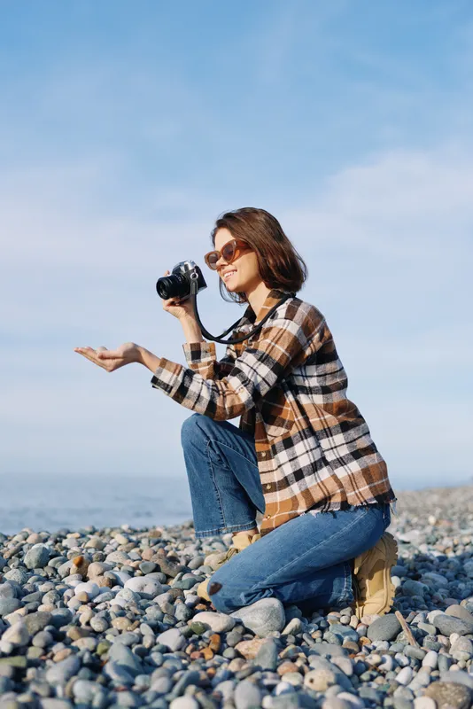 Photographer kneels on a rocky beach, smiling behind a DSLR, wearing a plaid shirt and jeans, capturing coastal scenes with natural light and a calm, expansive sky.