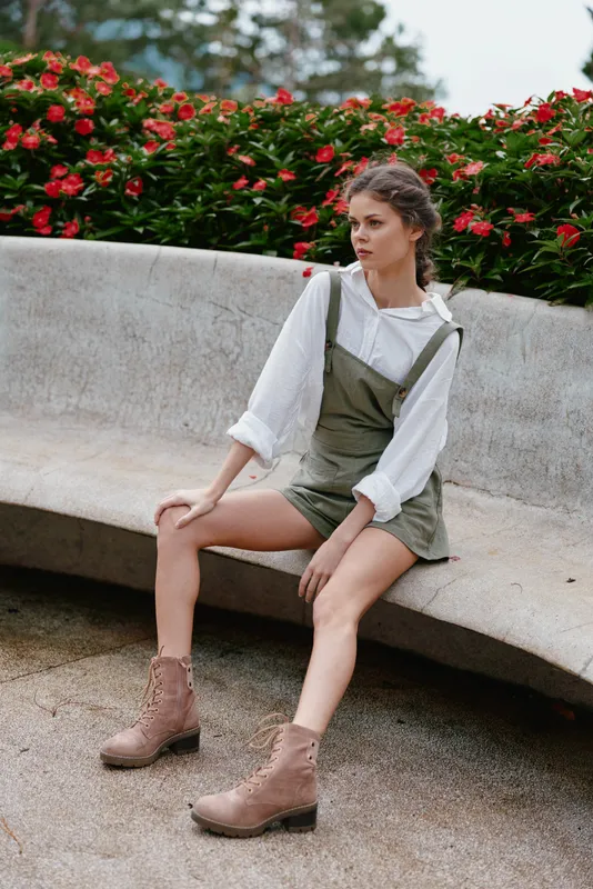 Young woman in stylish olive green overalls and white shirt sitting on a bench among vibrant red flowers, showcasing a casual and relaxed summer vibe
