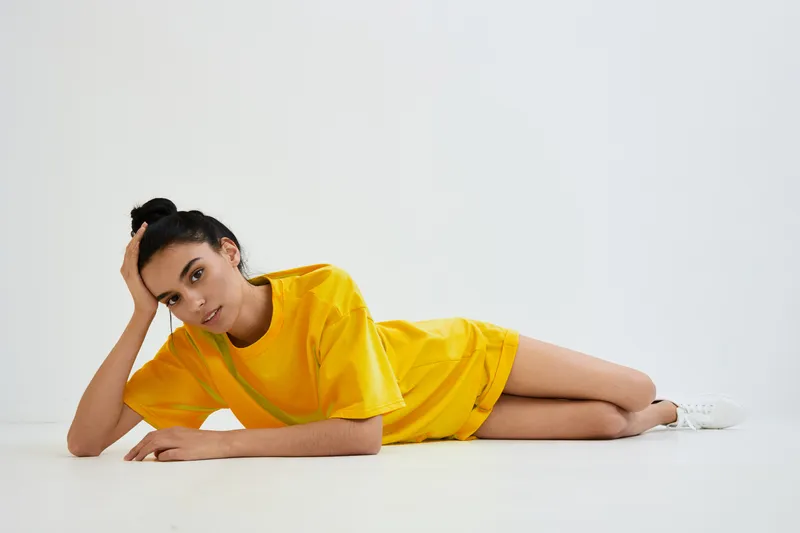 Young woman wearing yellow casual outfit lying on white floor with relaxed expression and black hair tied up in a bun, minimalistic bright studio setting.