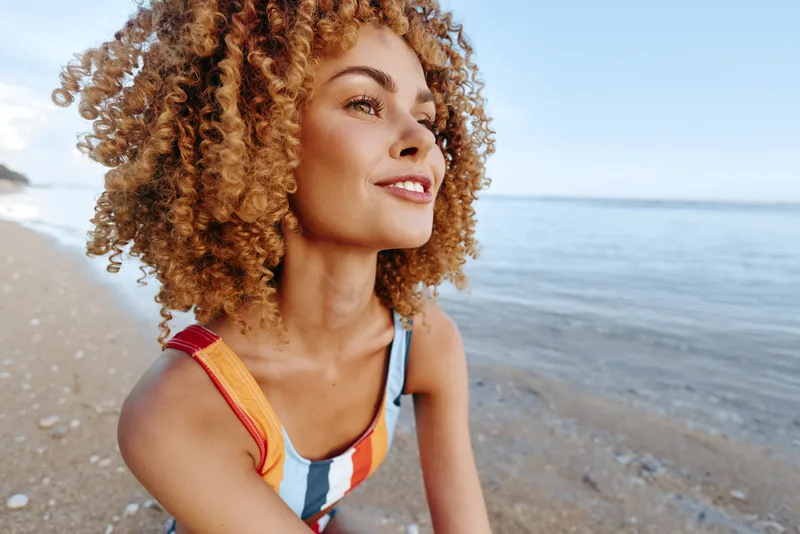 Beach portrait of a young woman with curly hair smiling in a colorful swimsuit, standing on a sandy shoreline with sunlit waves in the background and a gentle coastal breeze enhancing the carefree