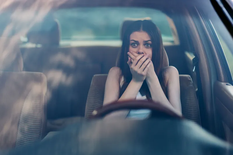 Young woman feeling anxious in a car, covering her mouth with hands, showing fear and stress while driving, with a blurred background and a warm color palette.