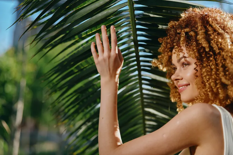 Smiling woman with curly hair enjoys sunny day outdoors, touching large green palm leaf, wearing white sleeveless top, natural light and vibrant background.