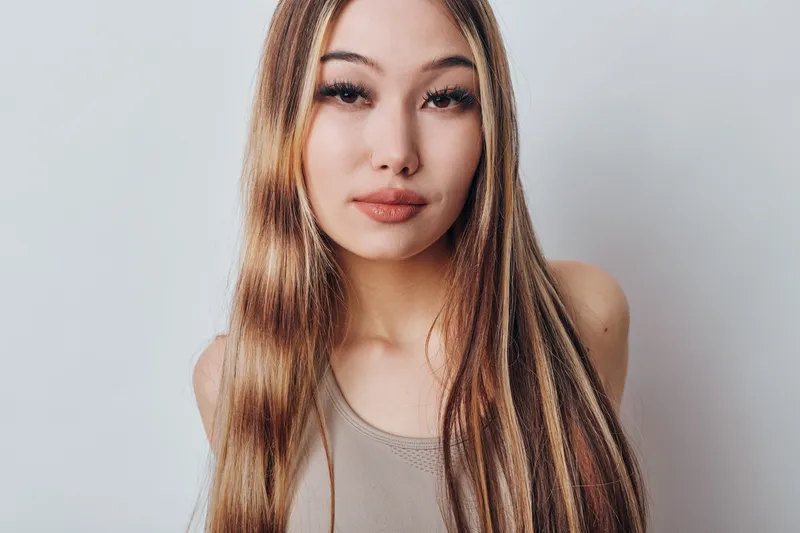 Young Asian woman with long straight hair wearing neutral top posing against white background in studio for beauty fashion portrait and natural look with minimal makeup.