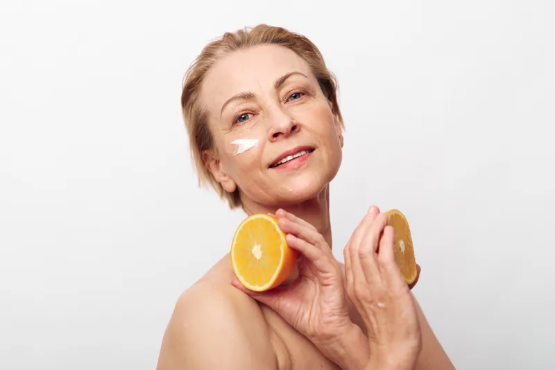 Mature woman with short hair holds two orange halves near face with cream on cheek. Bright studio background highlights natural skincare, healthy lifestyle, and citrus fruit for beauty and wellness.