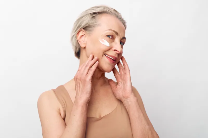 Smiling mature woman applies facial cream on clean skin with fingers, showing skincare routine and self-care concept. Studio portrait on white background with soft natural lighting.