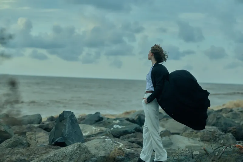 Windy shoreline, cape flowing, business attire, ocean horizon, rocky coast, dramatic pose, sea breeze, fashion style, solitary figure at the edge in moody light.