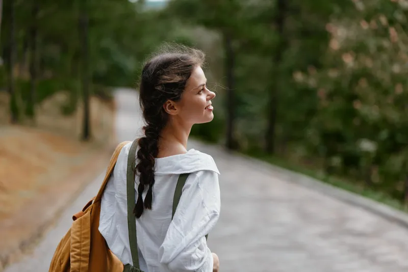 Young woman enjoying nature in a park, wearing a white shirt and carrying a brown bag, reflecting a peaceful and contemplative mood among greenery