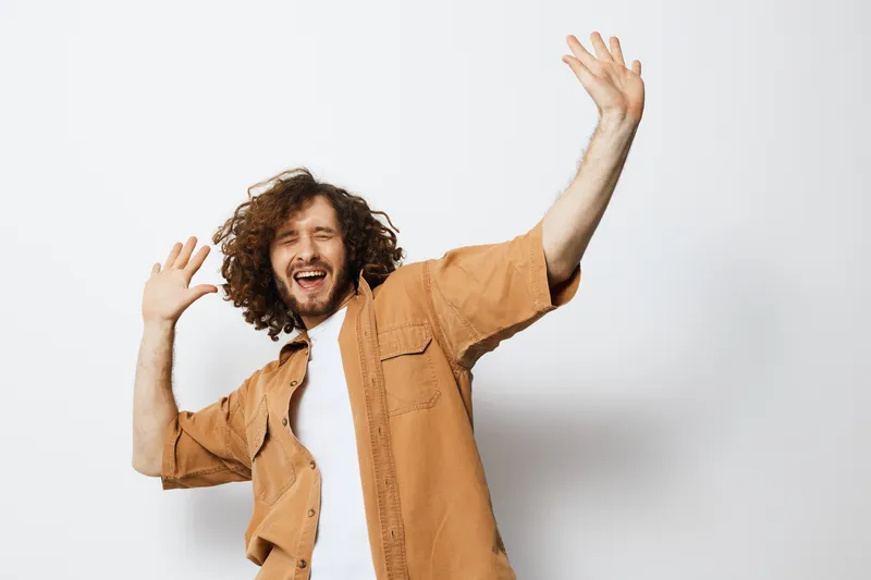 Joyful man celebrating with raised hands against a light background, wearing a casual tan shirt, embodying happiness and positive emotions