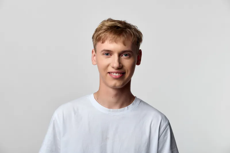 Young man wearing a white t shirt smiles softly in a clean studio setting, casual everyday style, bright lighting, relaxed mood, minimal backdrop, suitable for lifestyle or fashion content