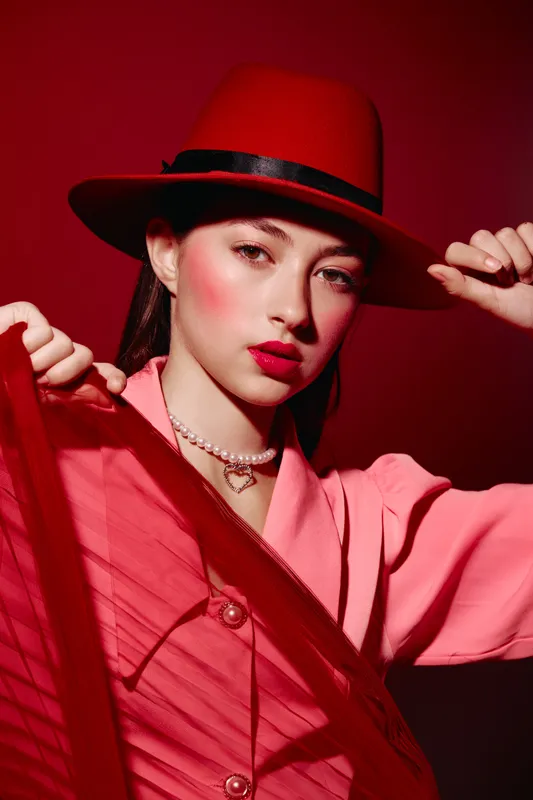 Young woman with red hat and bold makeup posing under red light. Creative beauty fashion photo with vibrant colors and elegant accessories, showcasing edgy, stylish look.