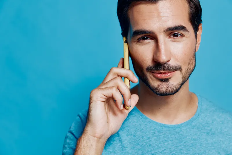 Smiling young man with a modern hairstyle wearing a blue t-shirt talking on a yellow smartphone against a vibrant blue background. Lifestyle and communication concept.