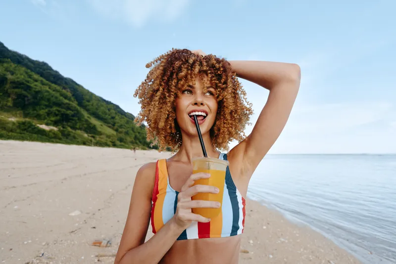 Young woman with curly hair enjoys a refreshing drink on a sandy beach near the calm sea on a clear sunny day, smiling and feeling relaxed in her colorful striped swimsuit.