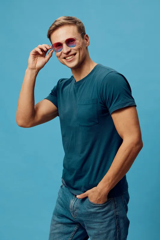 Smiling young man wearing blue t-shirt and pink sunglasses looks at camera, standing against plain teal background, casual pose. Vibrant, cheerful, summer lifestyle concept, fashion accessory.